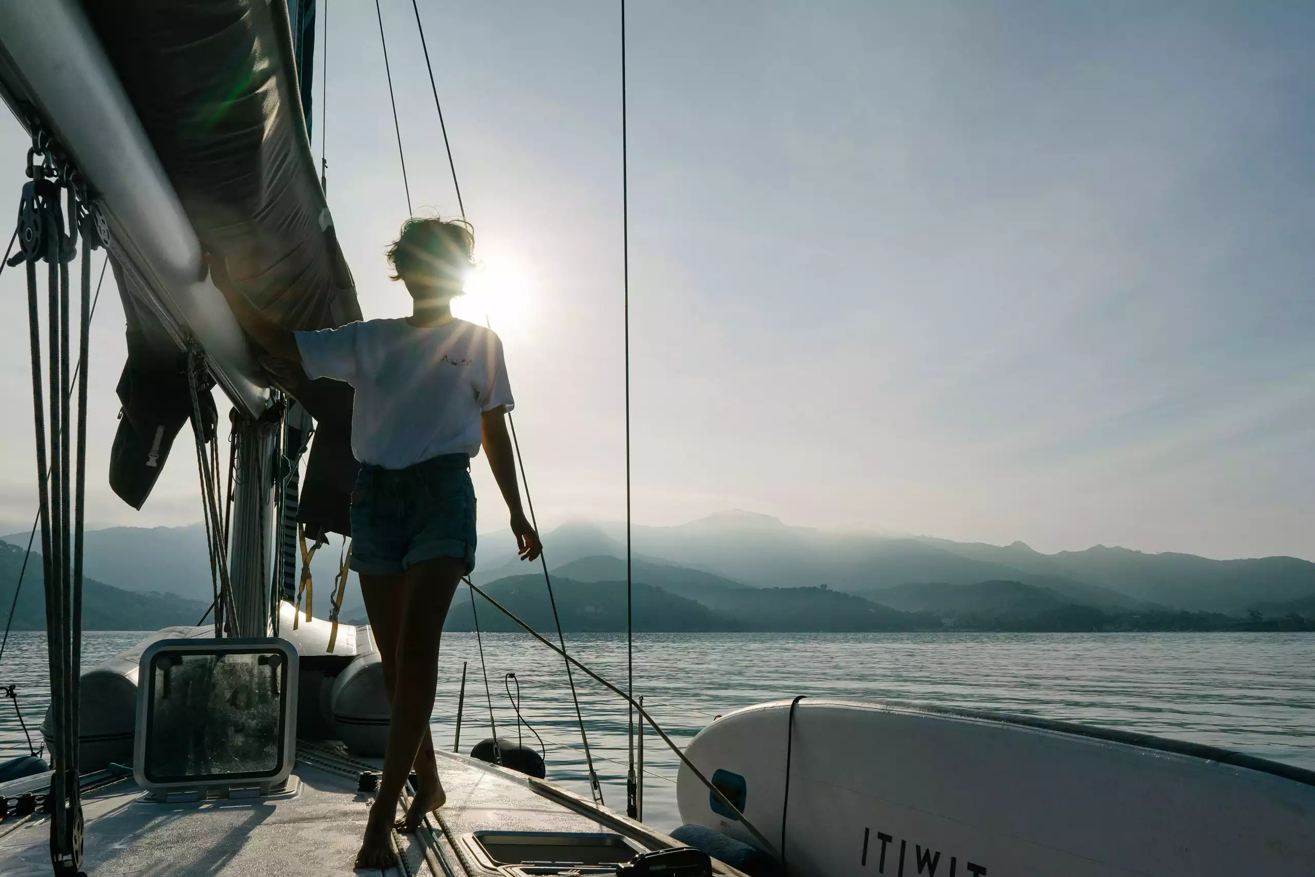 Silhouette einer Person auf dem Deck einer Yacht bei Sonnenuntergang mit ruhigem Wasser und Bergen im Hintergrund