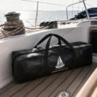 Schwarze Wobbeltasche mit Segel-Logo liegt auf dem Teakholzdeck einer Yacht neben Tauwerk und Winsch