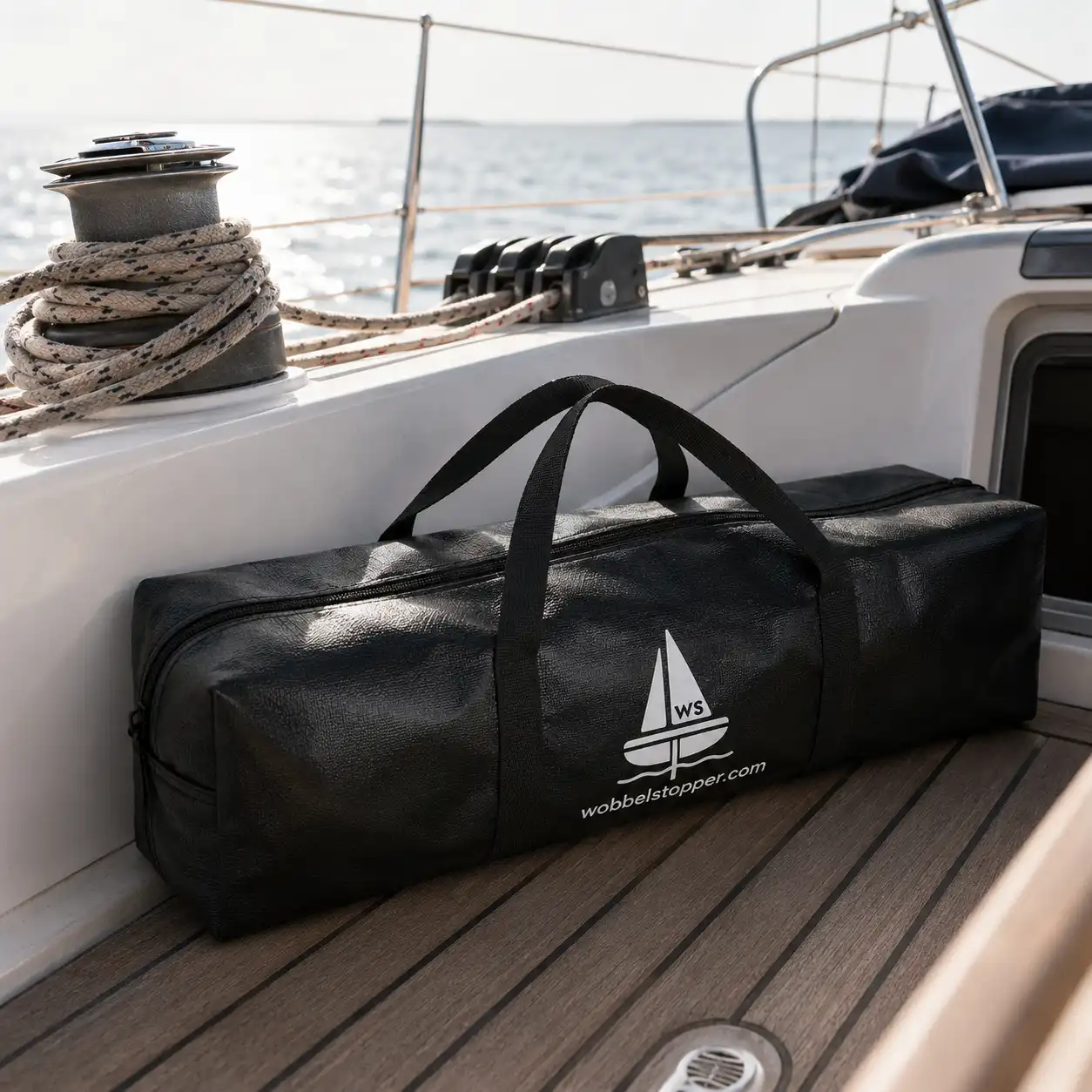 Schwarze Wobbeltasche mit Segel-Logo liegt auf dem Teakholzdeck einer Yacht neben Tauwerk und Winsch